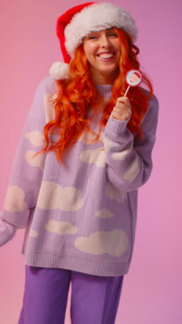 Vertical-Video-Studio-Portrait-Shot-Of-Young-Gen-Z-Woman-Wearing-Christmas-Santa-Hat-Holding-Candy-Lollipop-And-Dancing